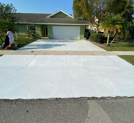 Fresh white concrete driveway leading to a green house with a garage and palm trees under a clear blue sky