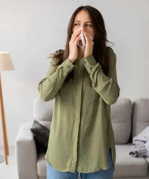 Woman in an olive shirt stands indoors, holding tissues to her nose in front of a sofa.