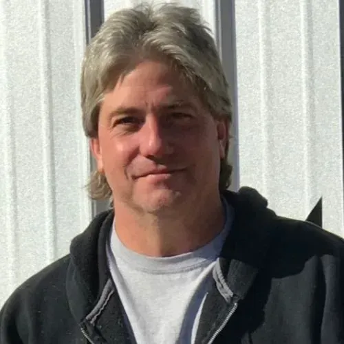 Man in a gray hoodie standing outdoors in front of a white metal wall, faintly smiling