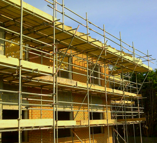 Scaffolding surrounds a brick building under construction, with multiple levels and wooden platforms.