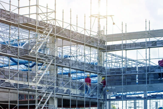 Construction workers on scaffolding at a building site, wearing hard hats and safety gear. Steel scaffolding surrounds a partially built structure.