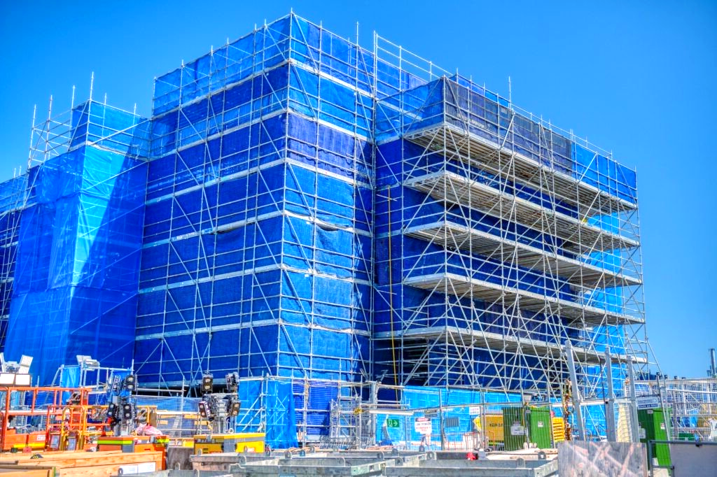 Construction site: blue safety netting covers a multi-story building with scaffolding against a bright blue sky.