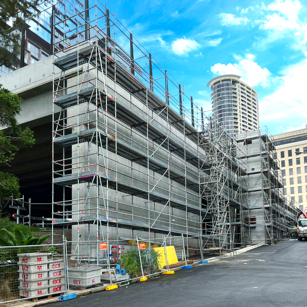 Scaffolding surrounds a building under construction, next to a road with a partially cloudy sky.