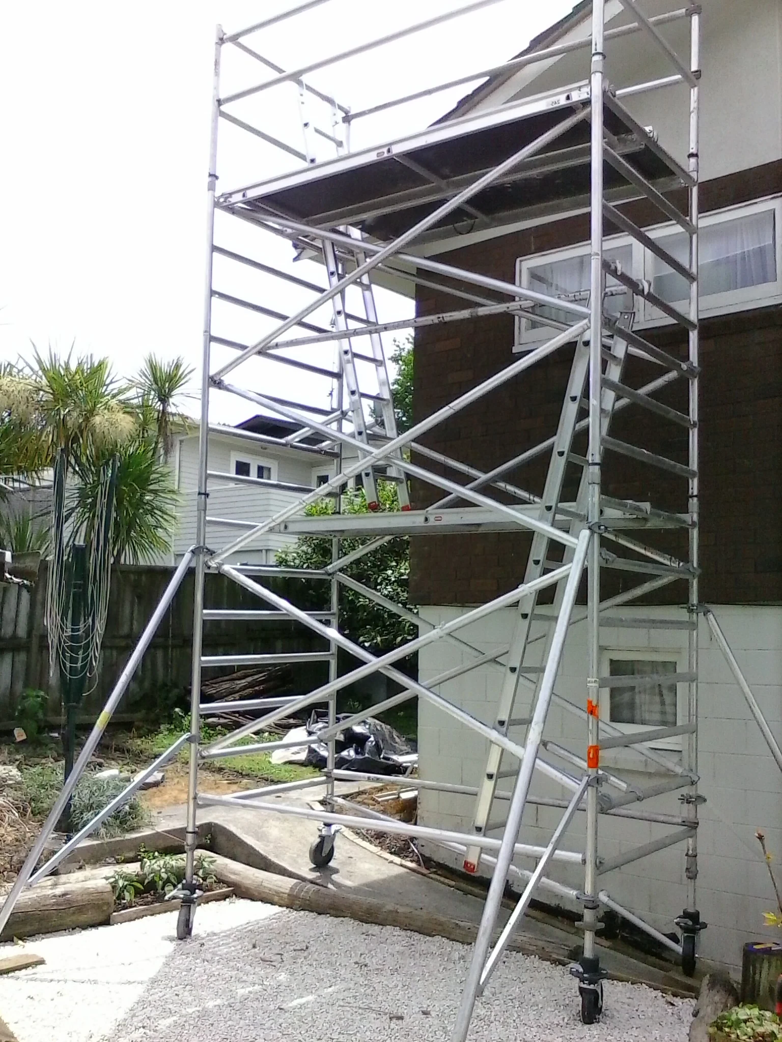 Aluminum scaffolding erected next to a two-storey building for exterior work. It sits on a gravel surface.