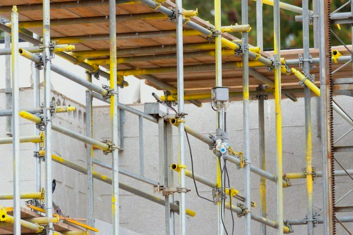 Scaffolding on a construction site, metal supports with yellow accents, against a white wall.