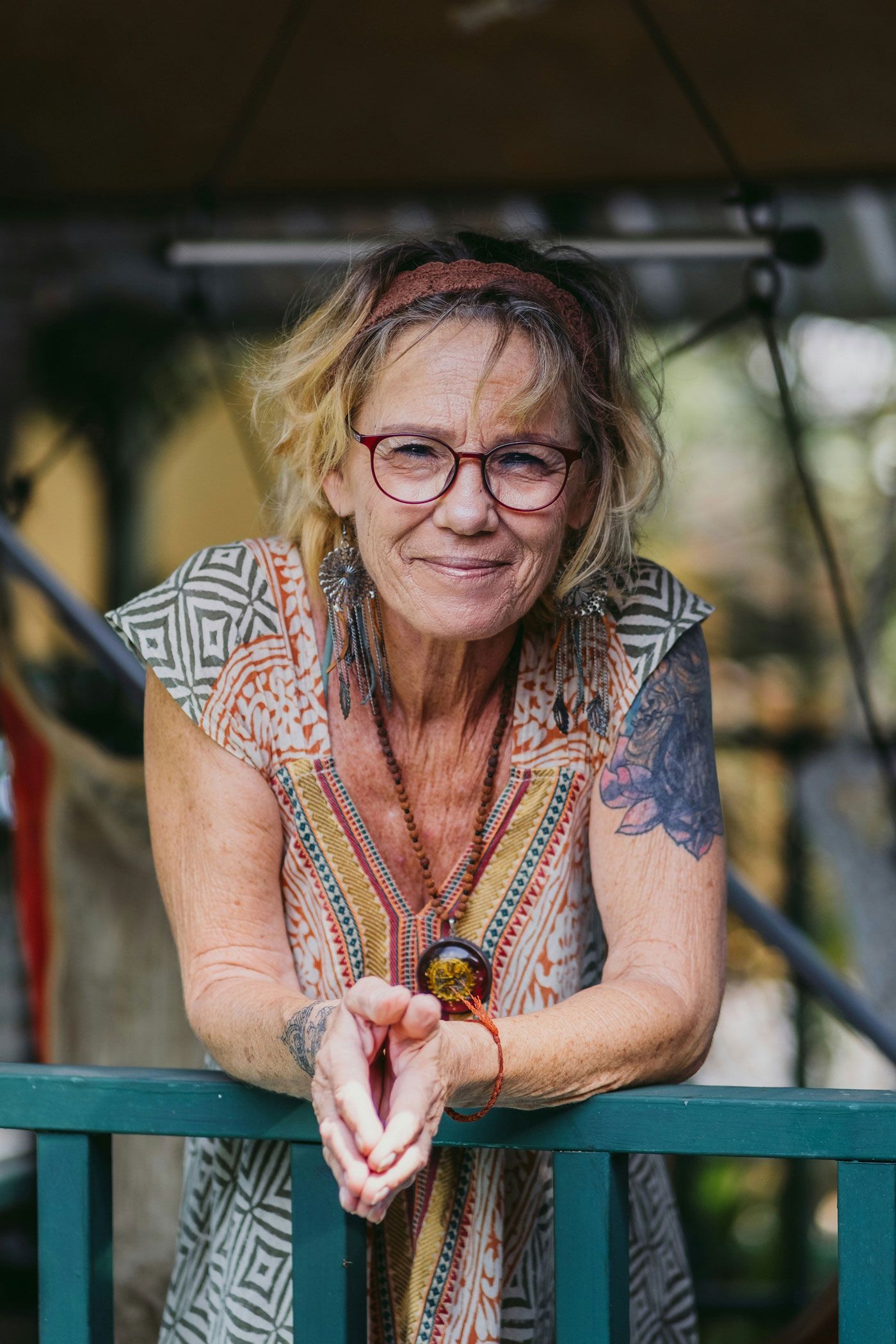 Person leaning on a teal railing, smiling outdoors with tattoos and glasses.