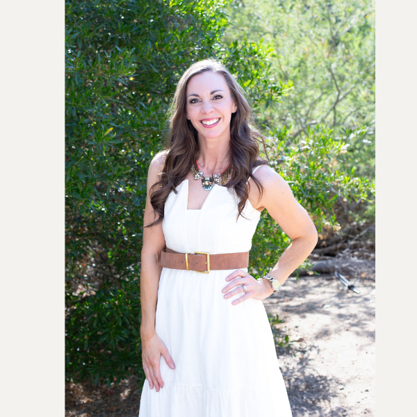 Woman in a white dress with a brown belt standing outdoors by greenery, smiling at the camera