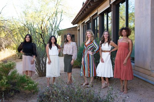 Five women in dresses standing outside a rustic building along a covered walkway.