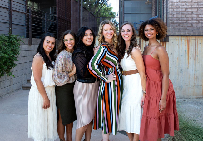 Six people posing outdoors in dresses, standing together and smiling near a fence.