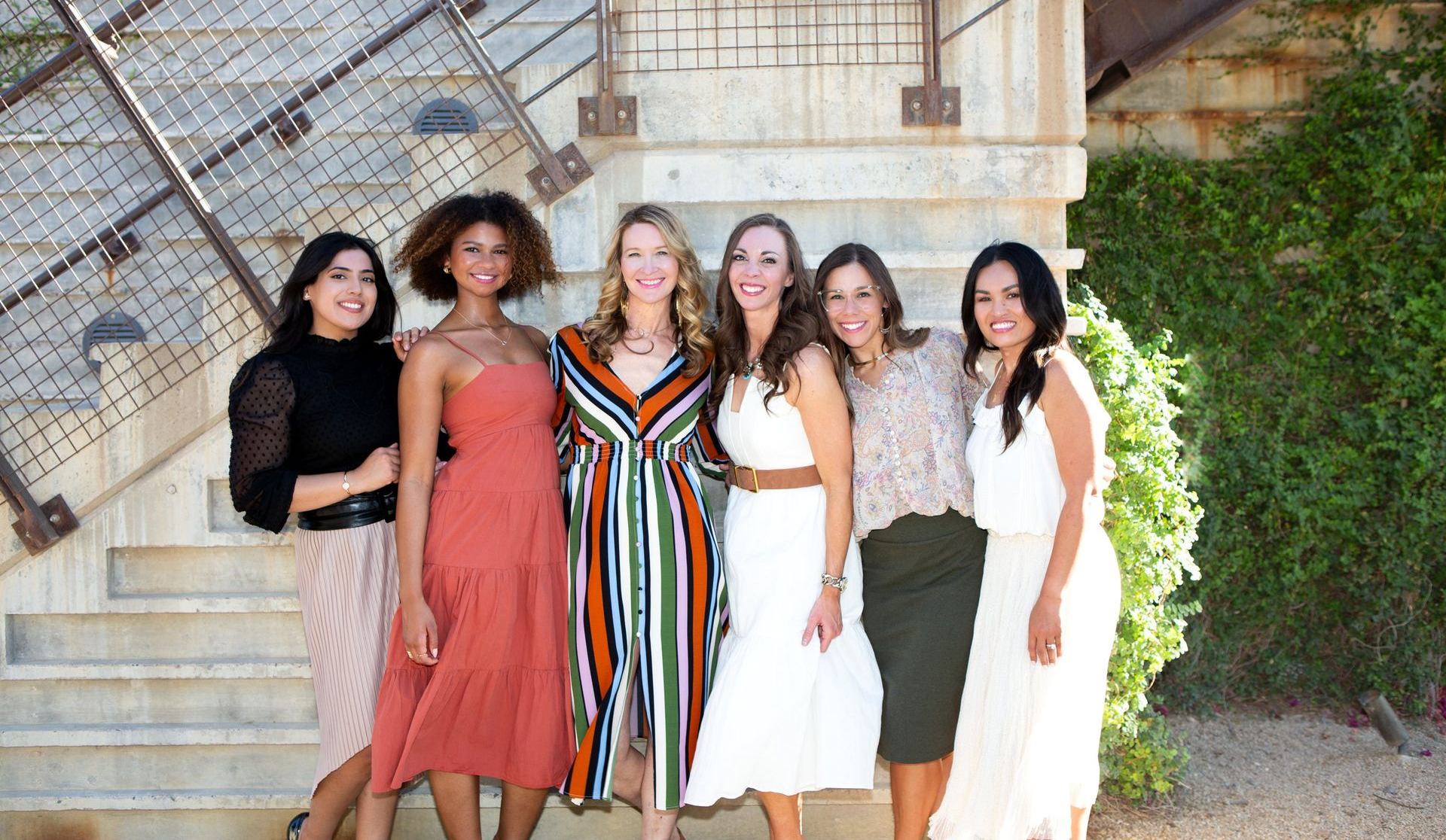 Six women standing together on outdoor steps, dressed in colorful outfits, smiling at the camera.