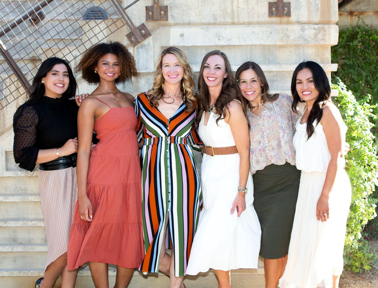 Six women standing together outdoors in colorful dresses, smiling at the camera.