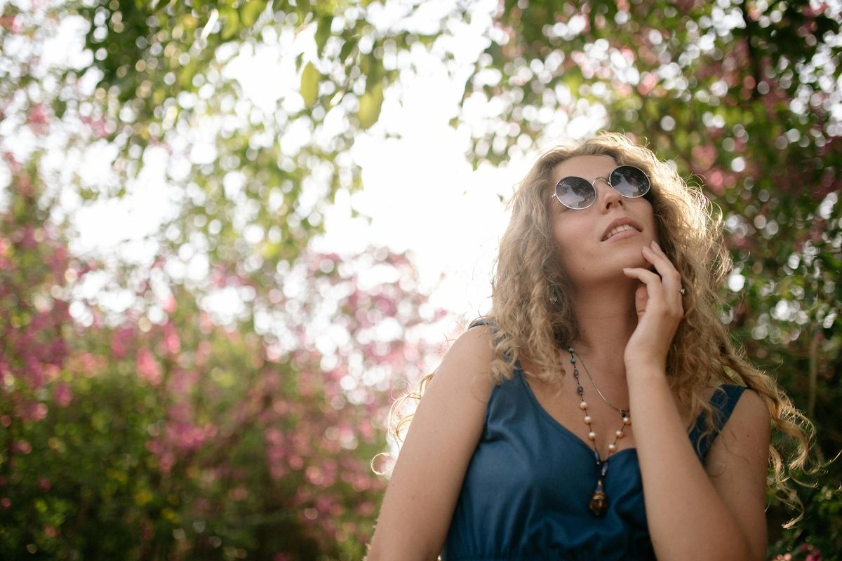 Woman in sunglasses gazes upward in a sunlit garden with pink blossoms.