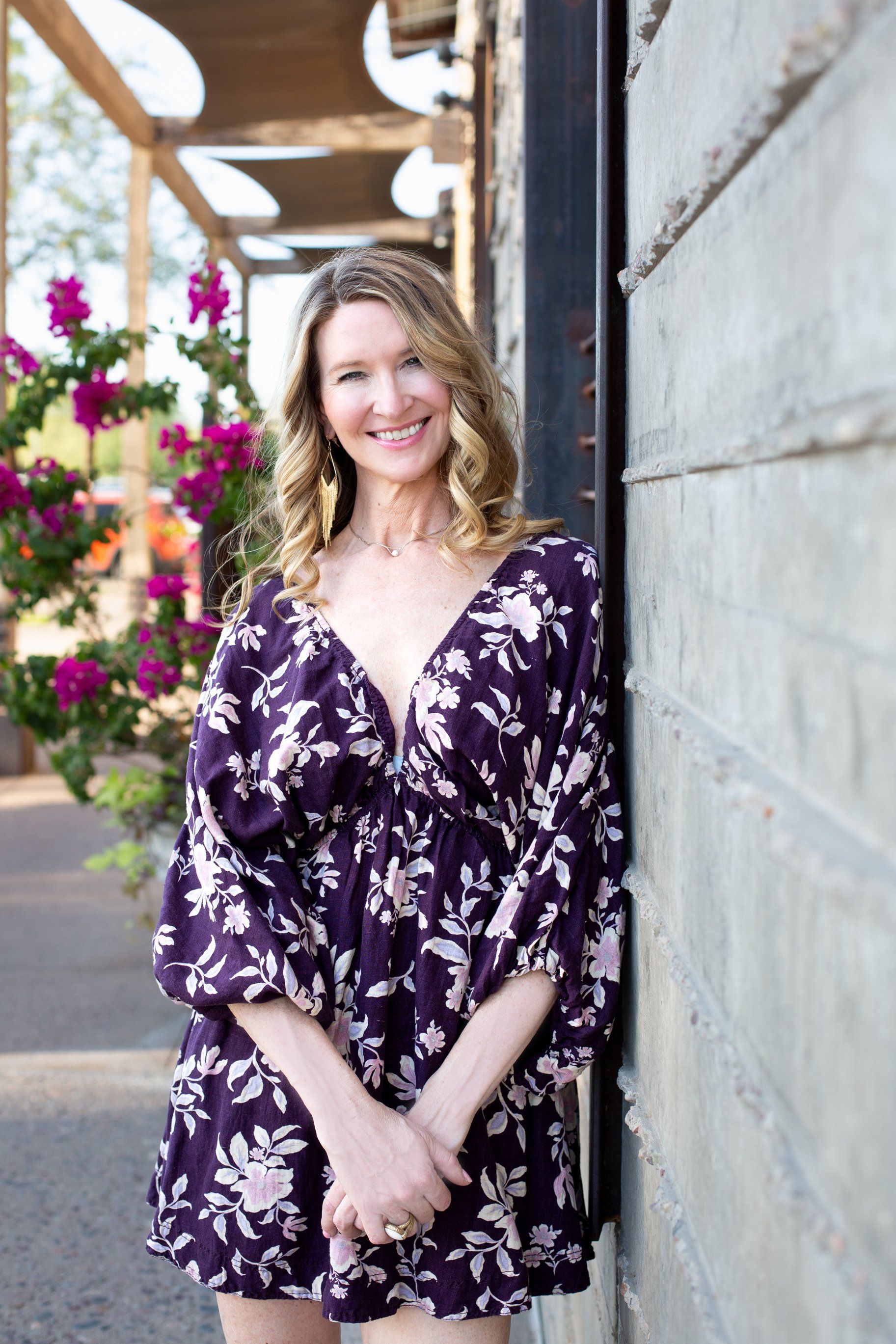Smiling woman in a purple floral dress leaning against a wall outdoors