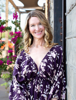 Smiling woman in a purple floral dress standing beside a wall outdoors