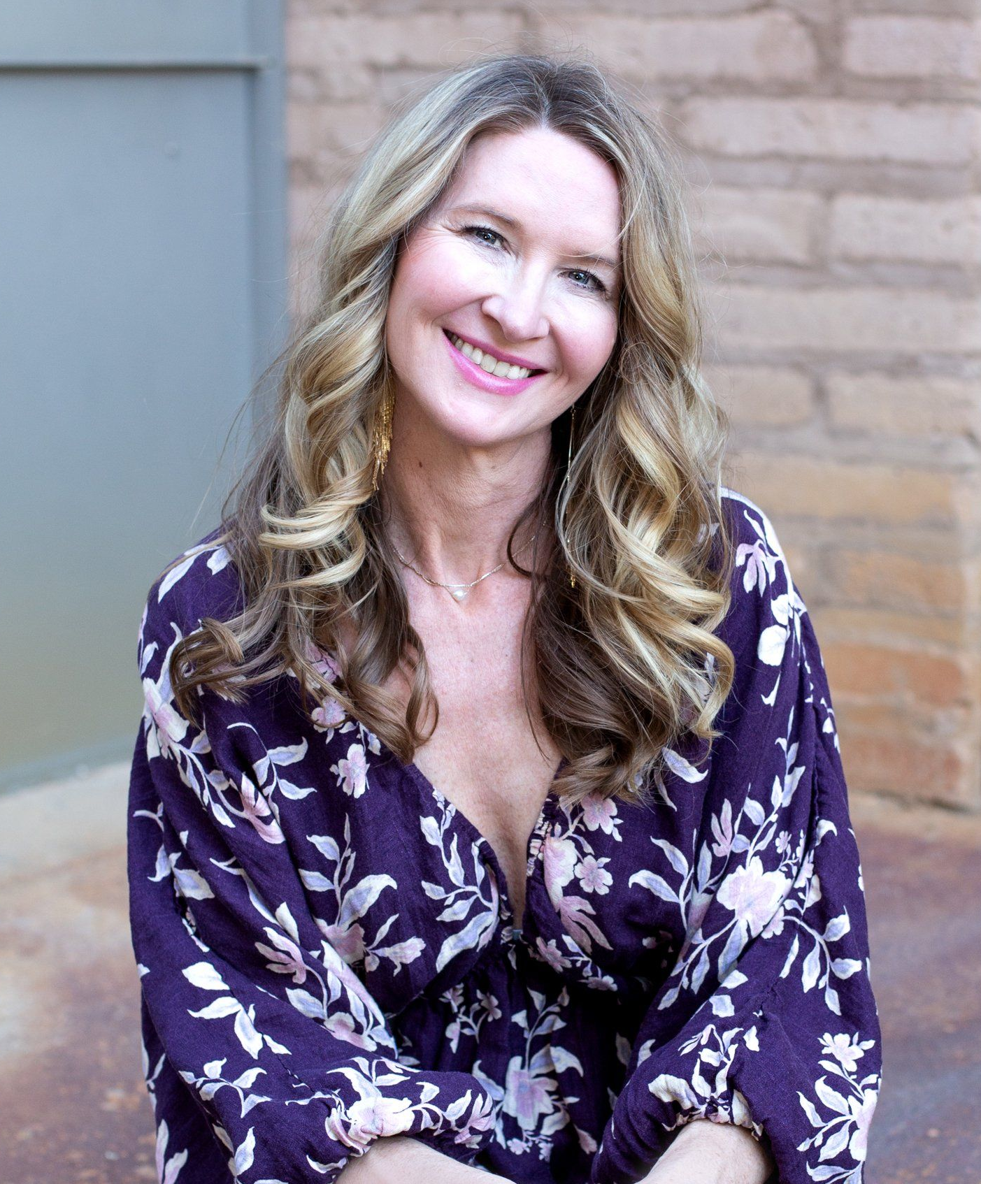 Smiling woman in a purple floral dress sitting against a brick wall