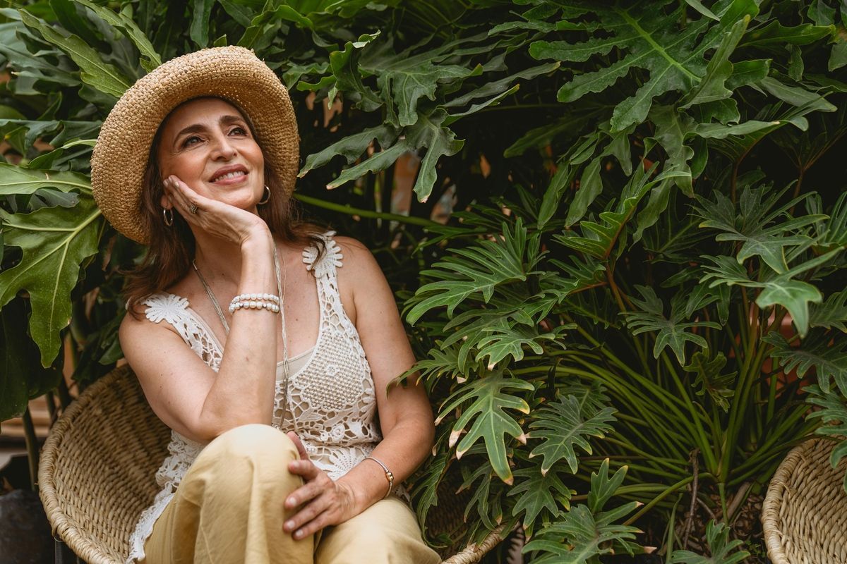 Woman in a straw hat sitting among lush green ferns, looking up thoughtfully