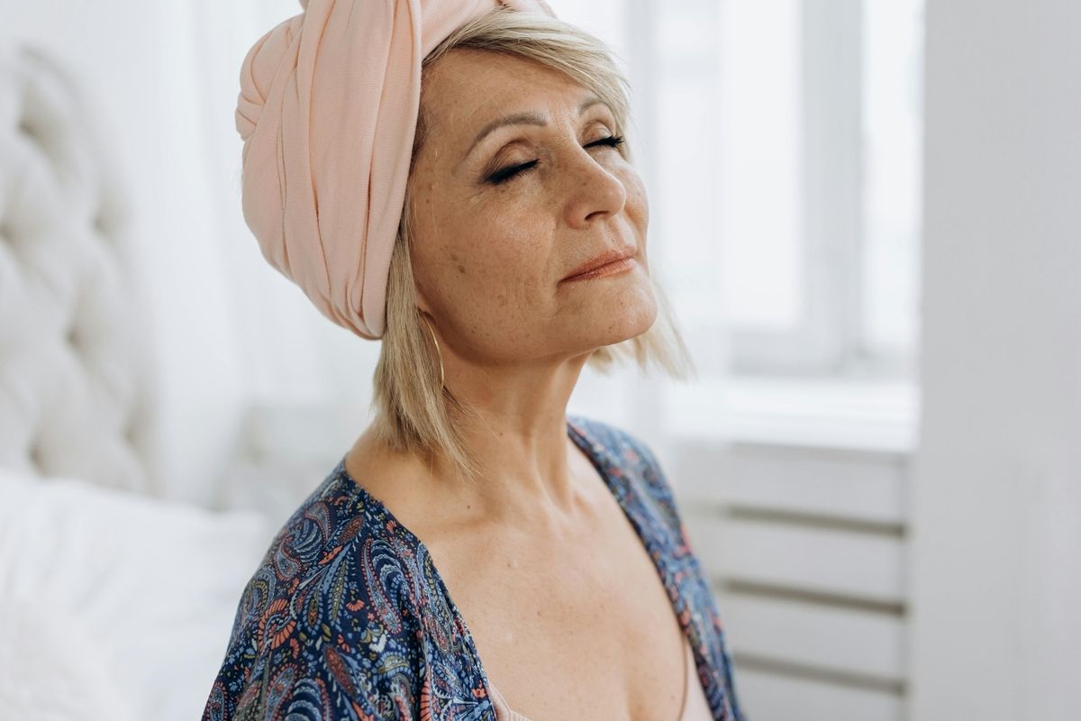 Woman with a pink headwrap sitting by a bright window, gazing upward with a calm expression