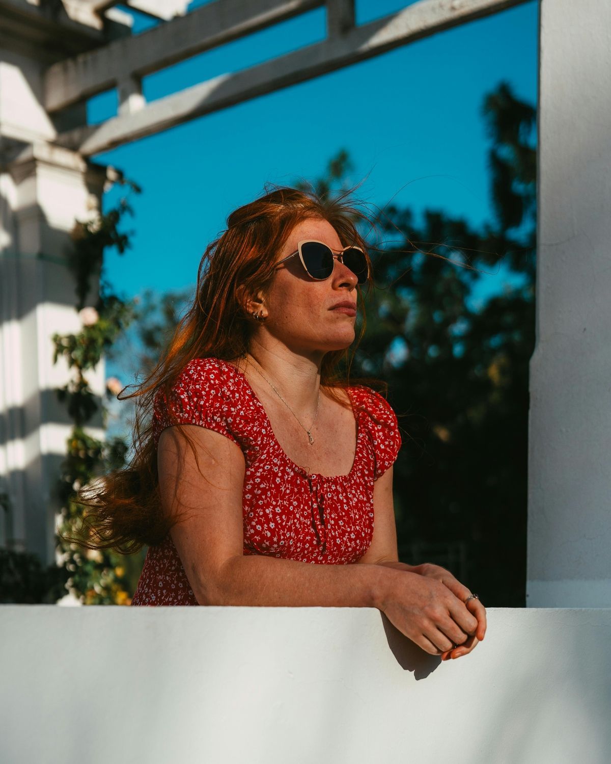 Woman in a red dress and sunglasses leaning on a white wall outdoors in sunlight