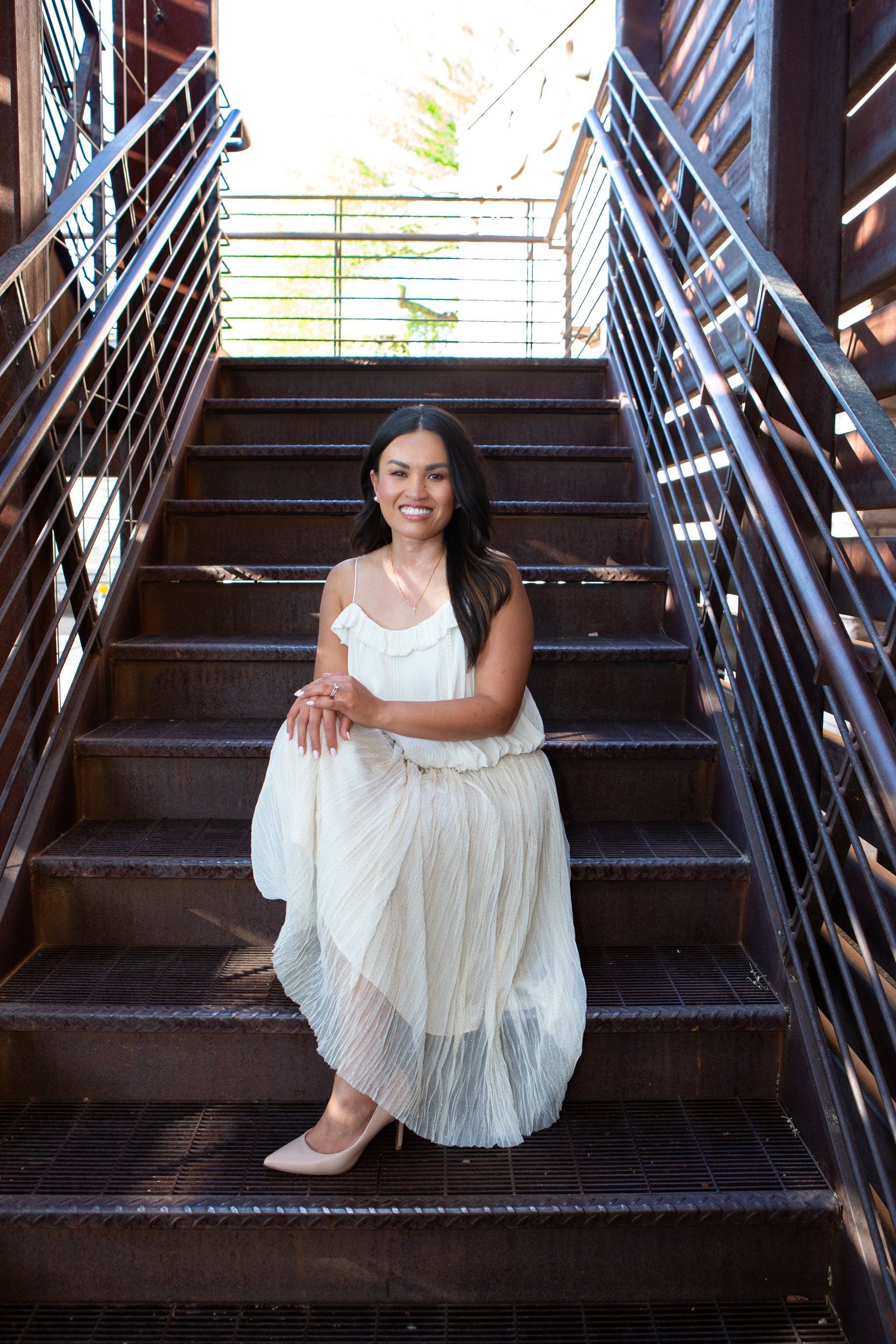 Woman in a white dress sitting on wooden stairs, framed by metal railings and bright daylight above.