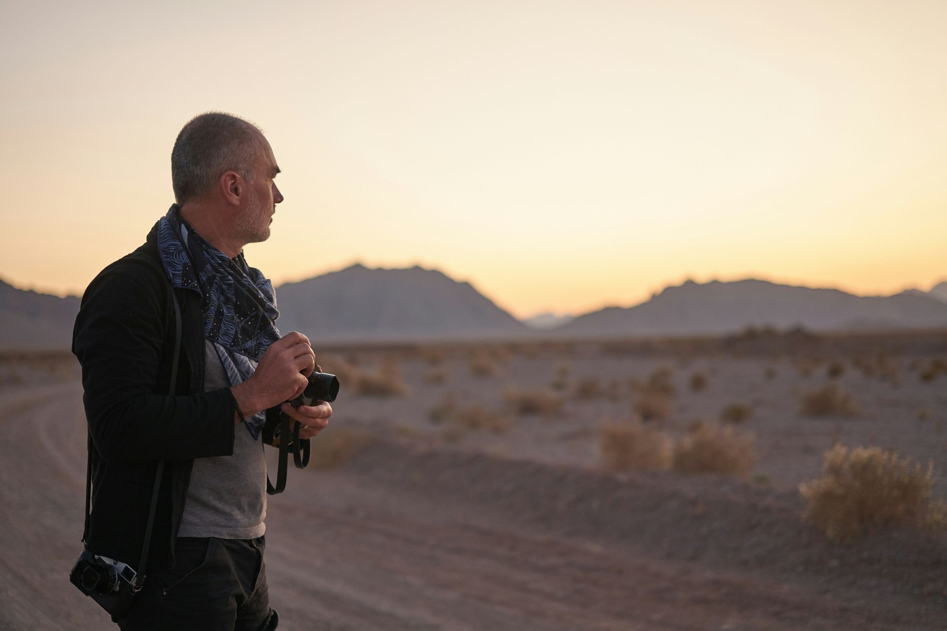 Man holding a camera at sunset in a desert landscape with mountains in the distance