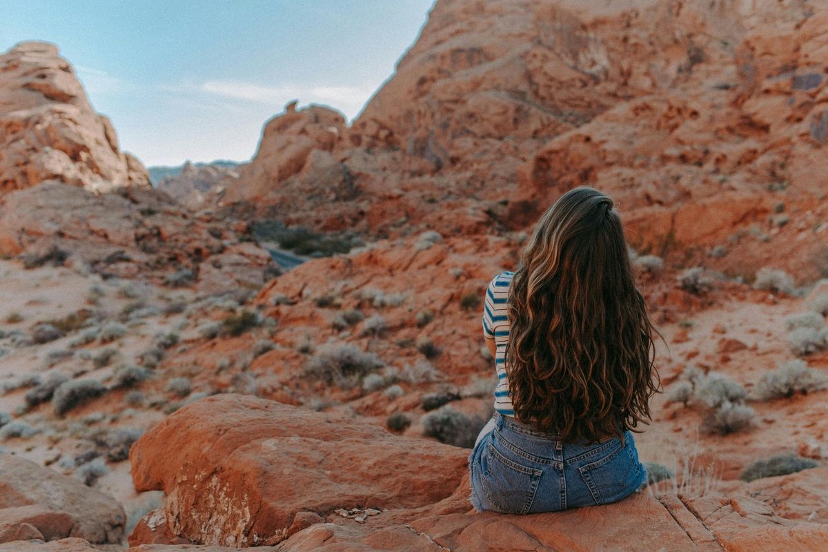 Person sitting on a rocky desert landscape, facing rust-colored hills under a blue sky.