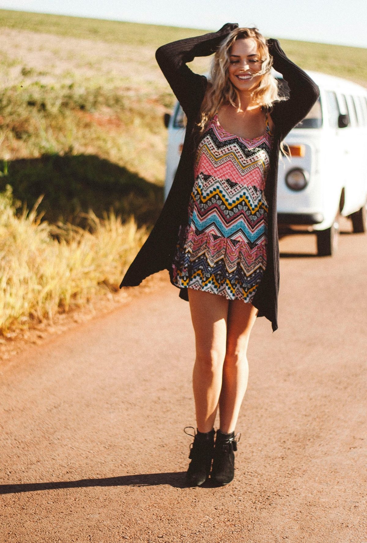 Woman standing on a road, smiling with hands in her hair, wearing a colorful dress and black cardigan.