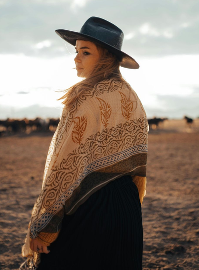 Person in a black hat and patterned shawl standing on a beach at sunset.
