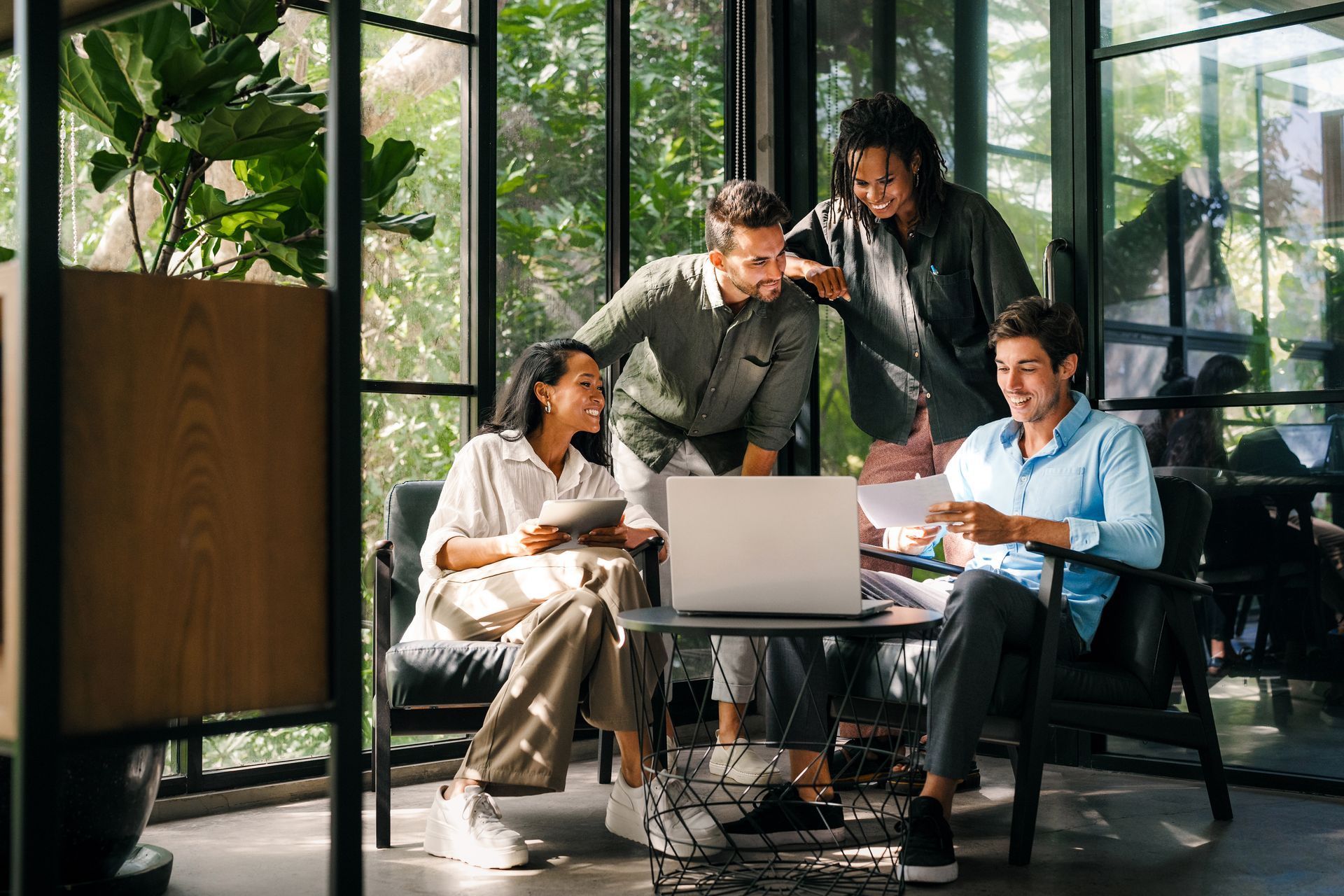 Four people collaborating around a laptop in a bright office with large windows overlooking greenery.