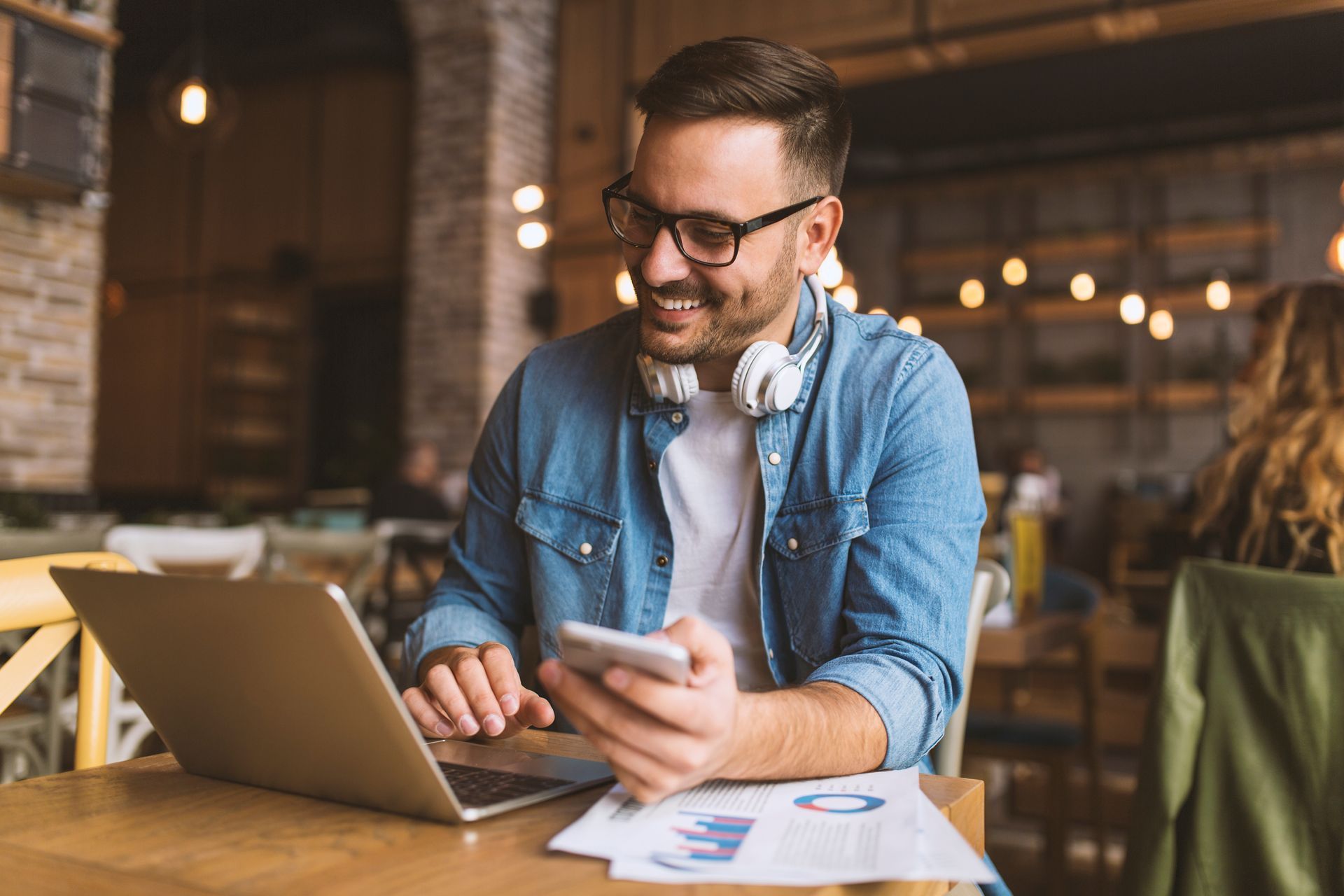 Man in glasses, smiling, uses a laptop and smartphone at a cafe table with papers.