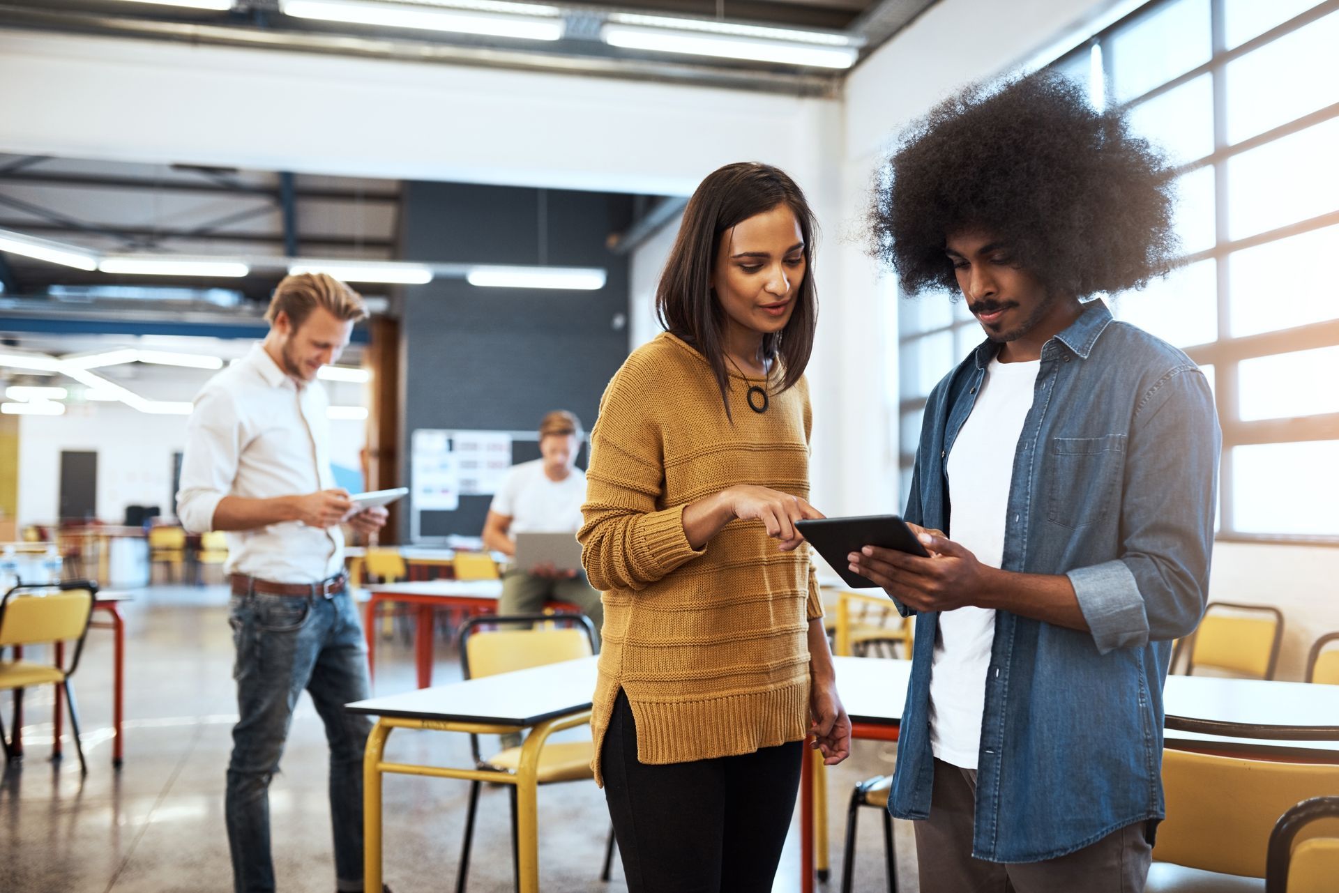 Two people looking at tablet, others working in modern office setting.