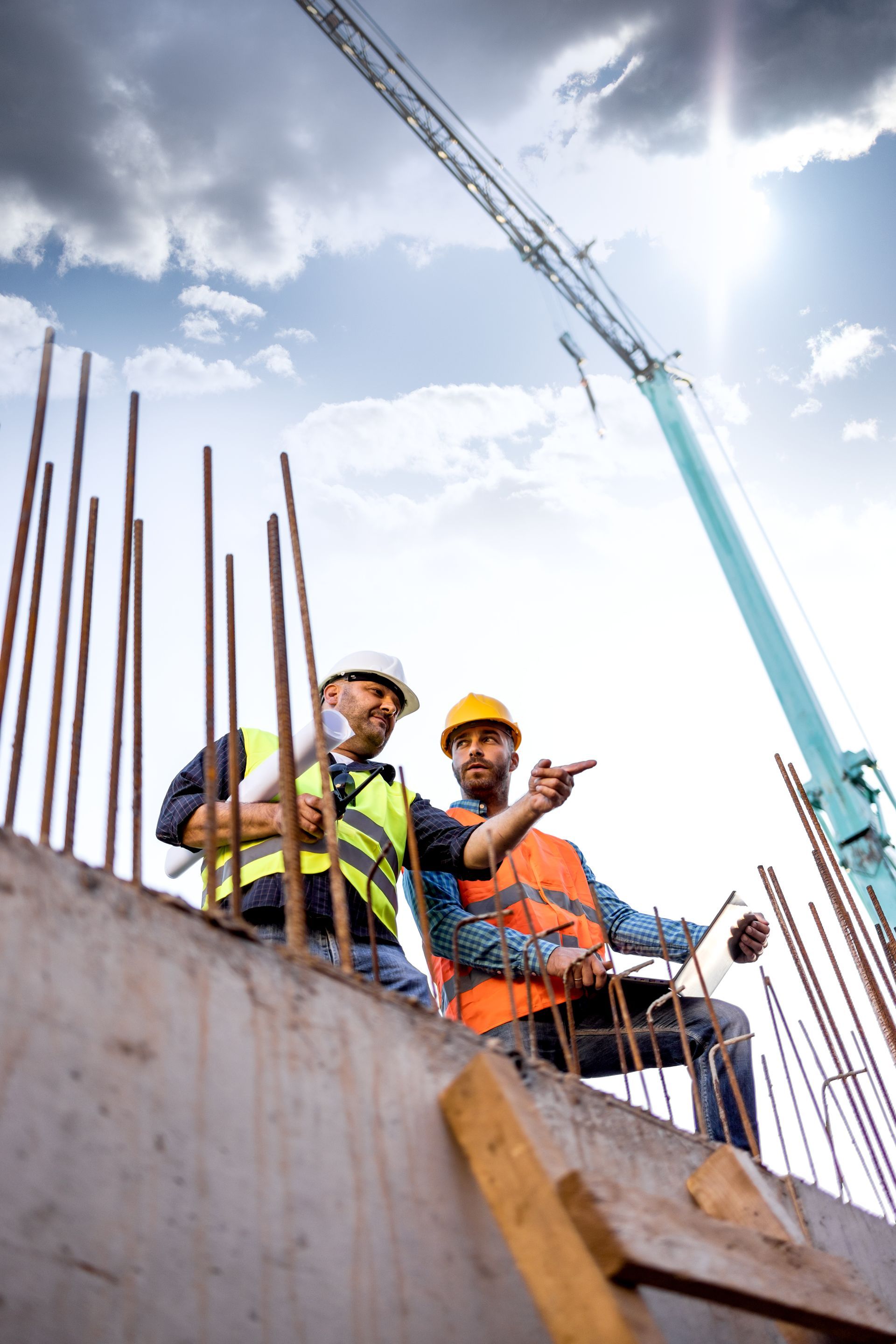 Two construction workers in safety vests on a concrete structure, discussing a plan, with a crane in the background.
