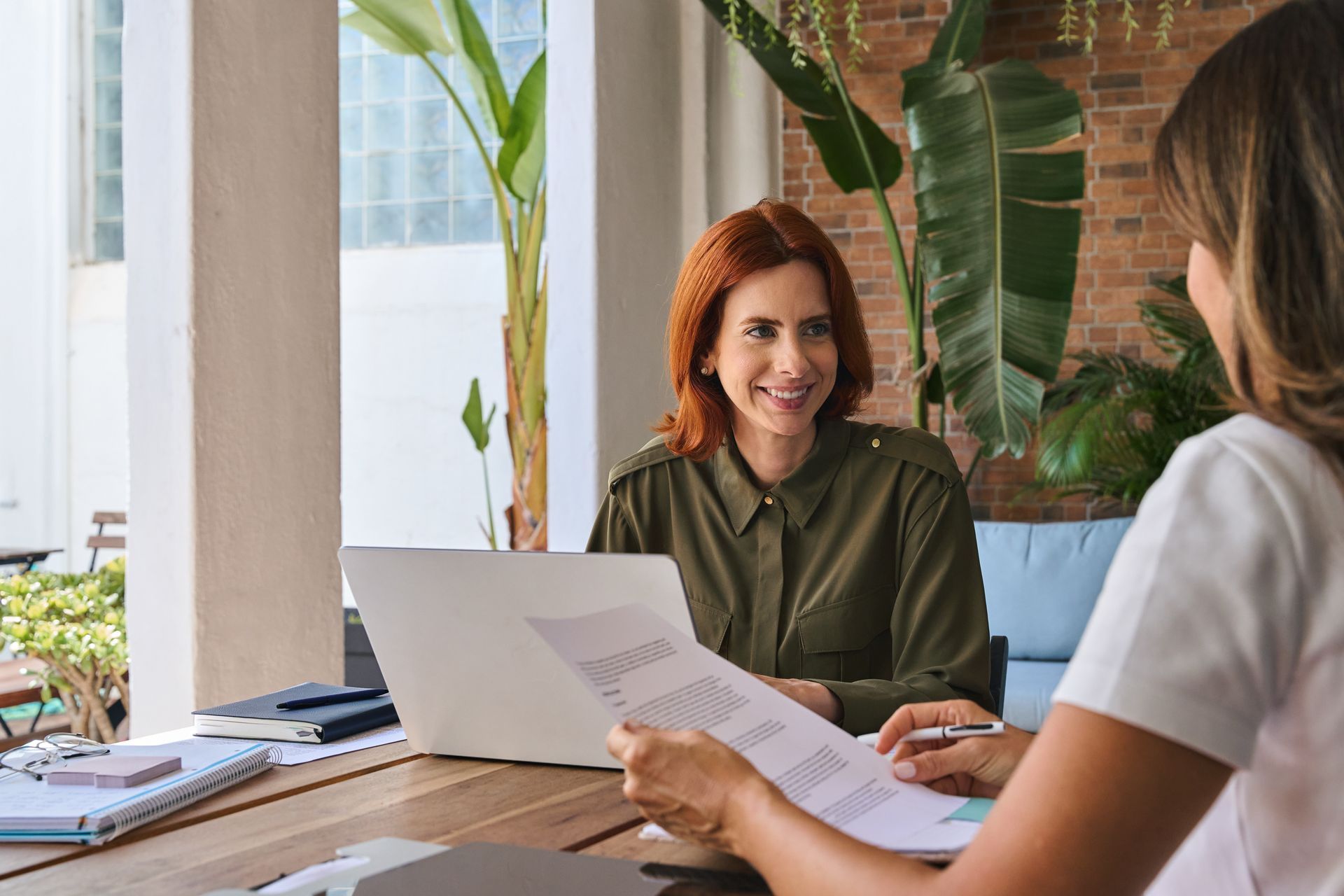 Woman with red hair smiling, seated at a table with another person, reviewing documents with a laptop present.