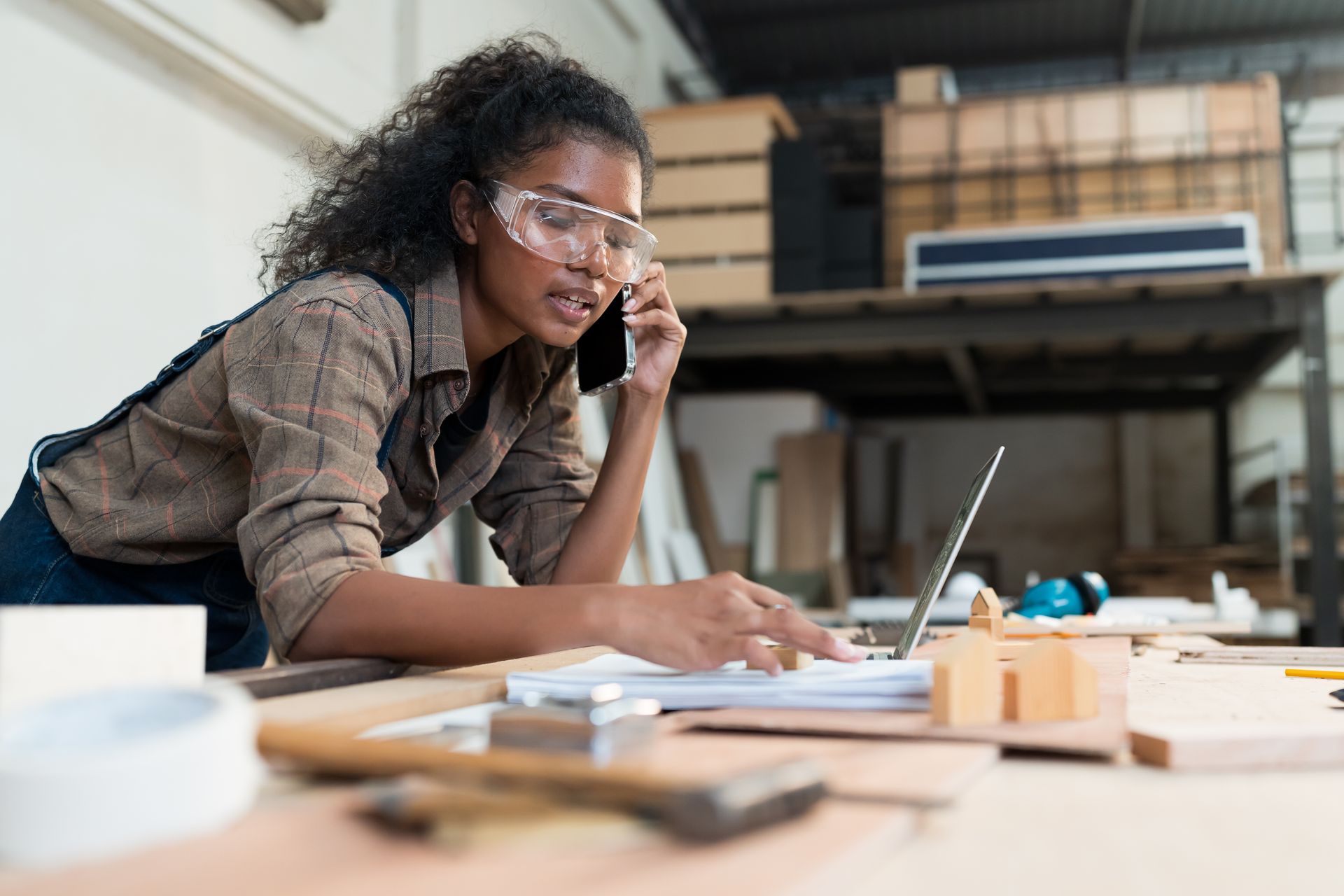 Woman in safety glasses uses laptop and phone in a workshop.