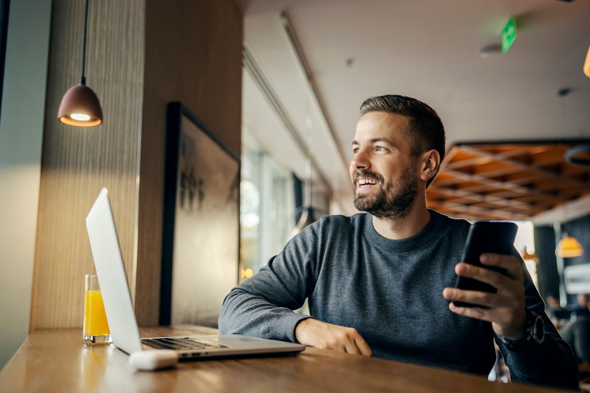 Man holding a phone, smiling, sitting at a table with a laptop, looking out a window in a cafe.
