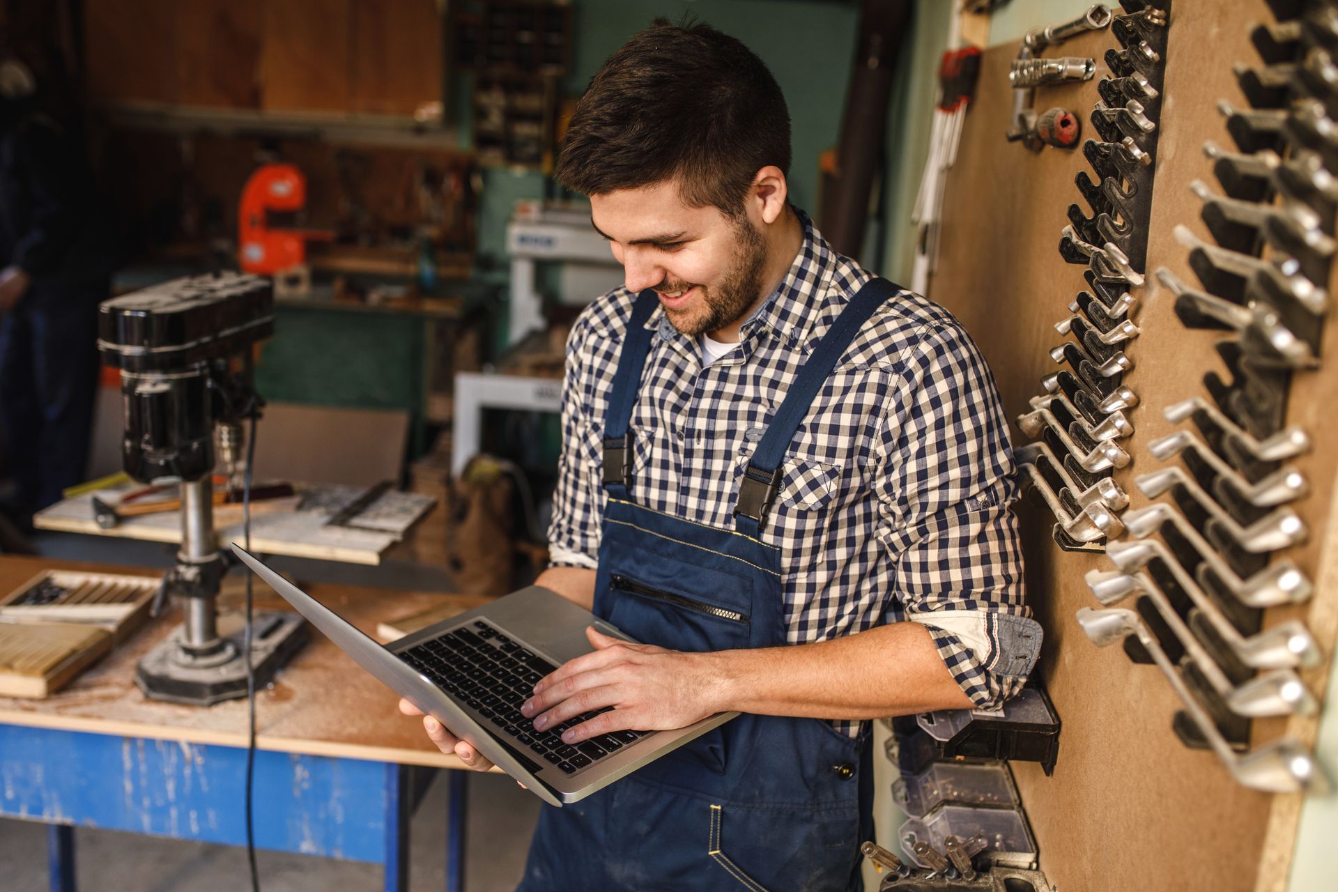 Man in overalls using a laptop in a workshop, smiling. Tools and drill press in background.