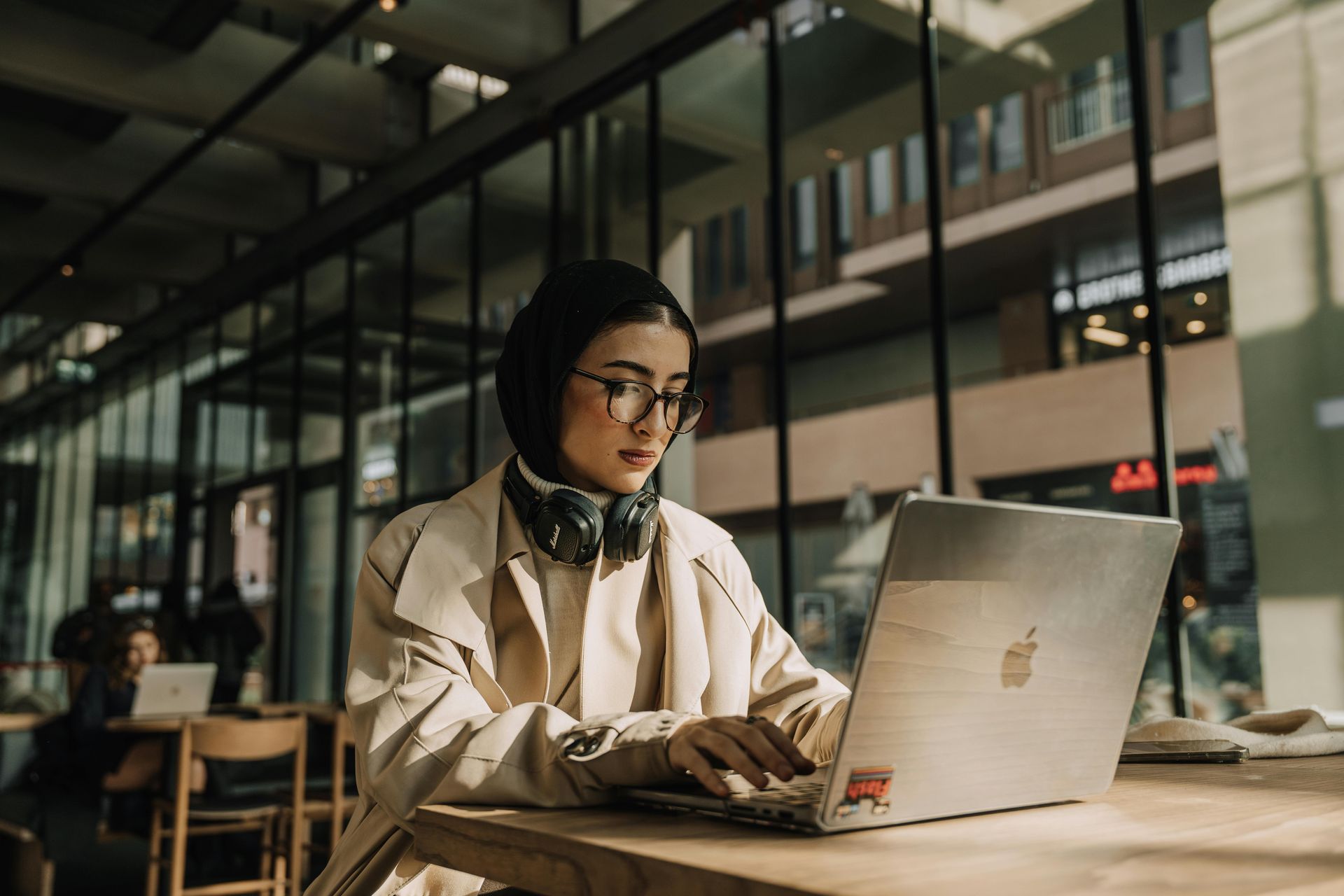 Woman wearing headphones and glasses, working on a laptop at a cafe table. Sunlight streams through a window.