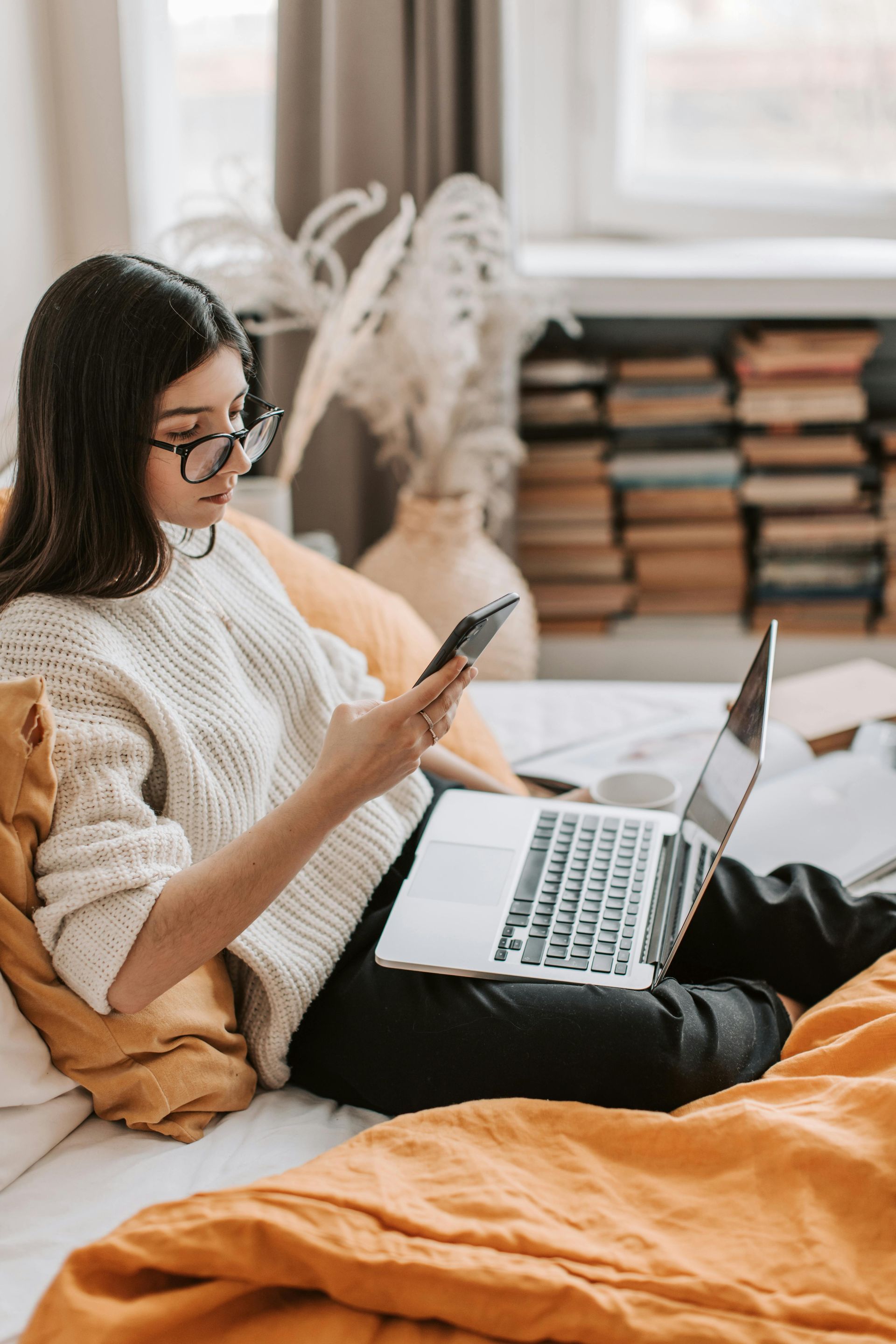 Woman with glasses using phone and laptop on a bed with orange bedding, near books.