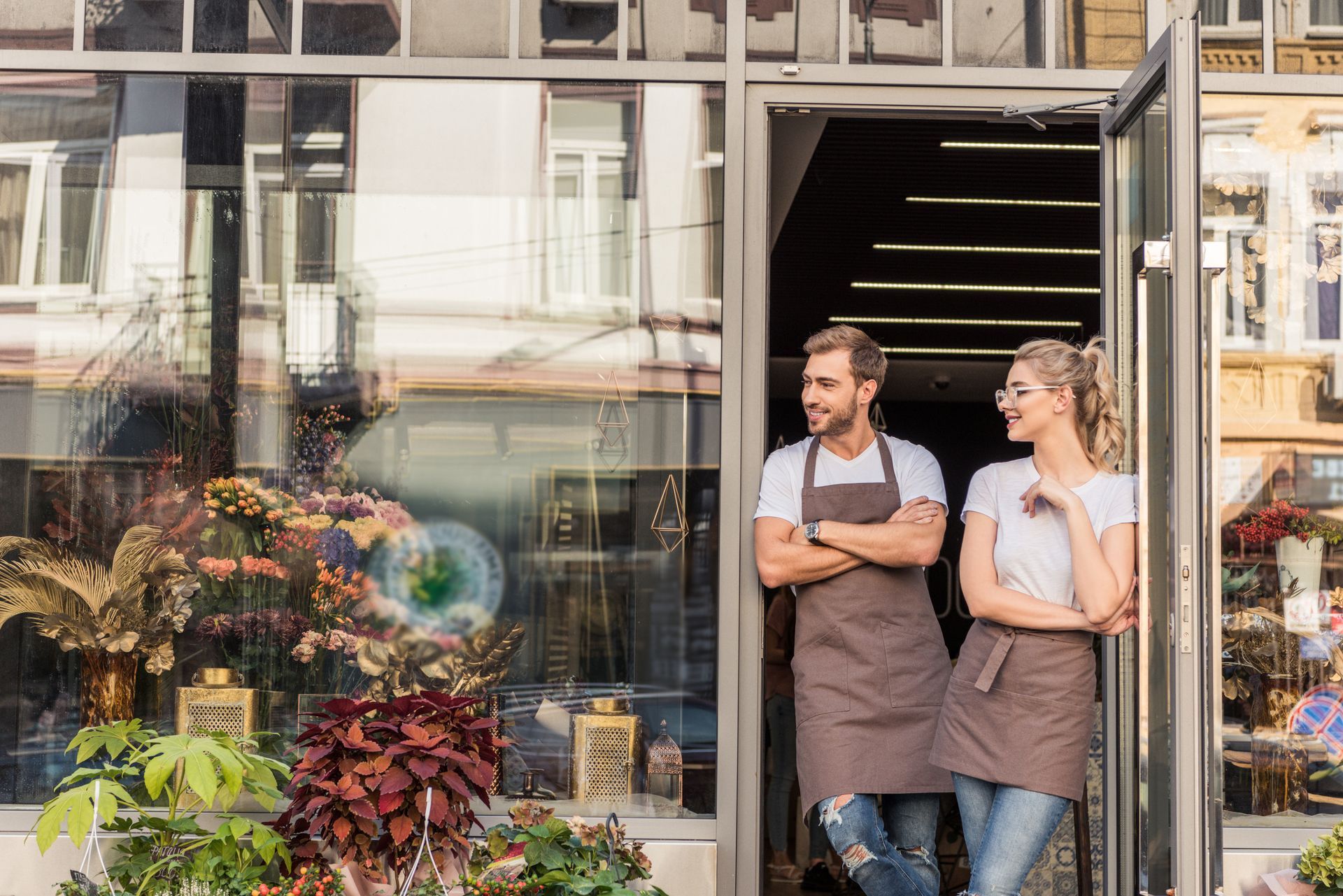 Two people in aprons leaning in a doorway of a shop, arms crossed, smiling. Shop window displays plants.