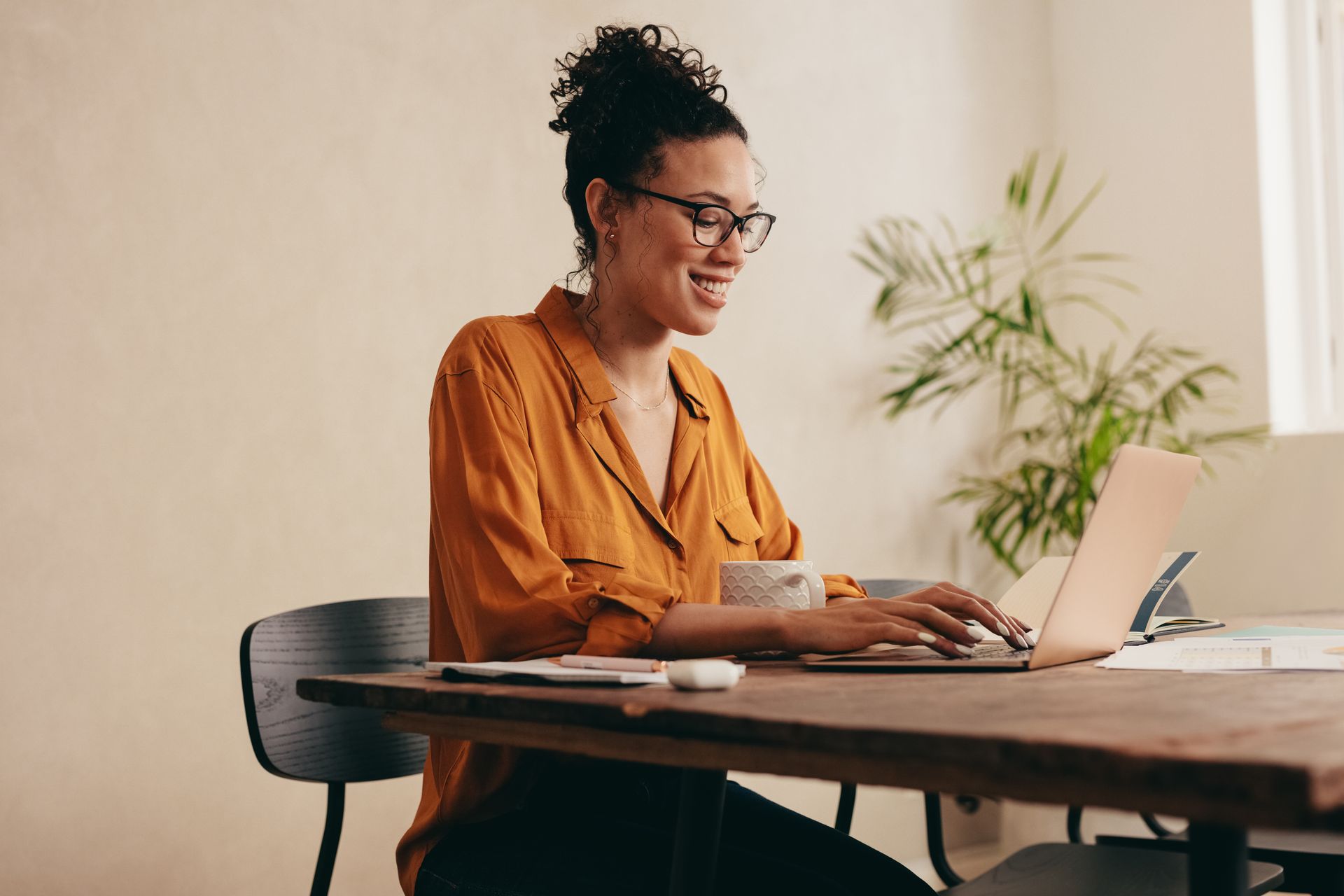 Woman in glasses, orange top, typing on a laptop at a wooden table, smiling, with a coffee mug and plant.