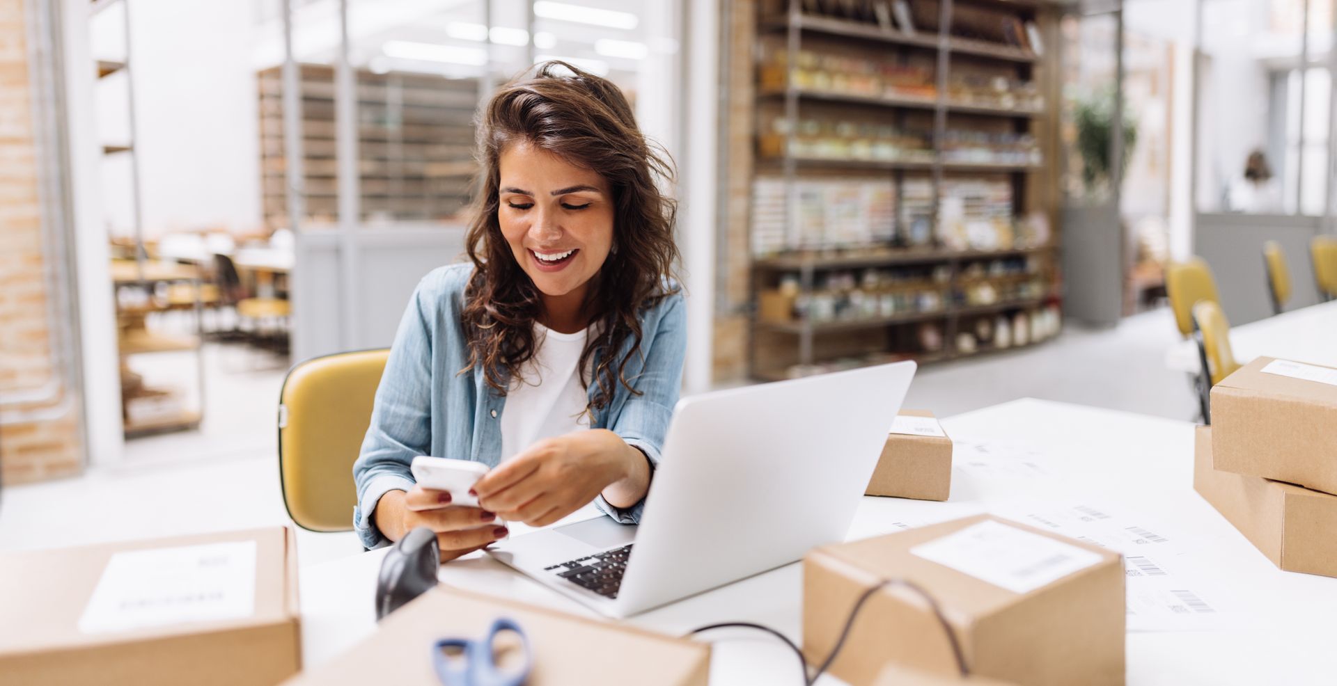 Woman smiles while using her phone and laptop, surrounded by shipping boxes in a bright workspace.