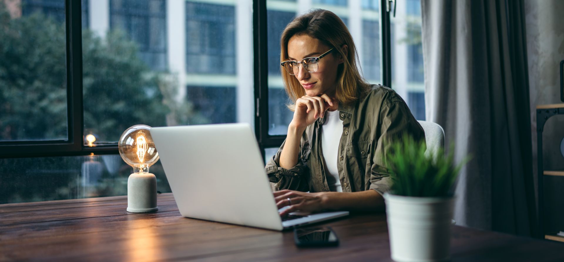 Woman with glasses using a laptop at a desk near a window. Desk includes a lamp and plant.