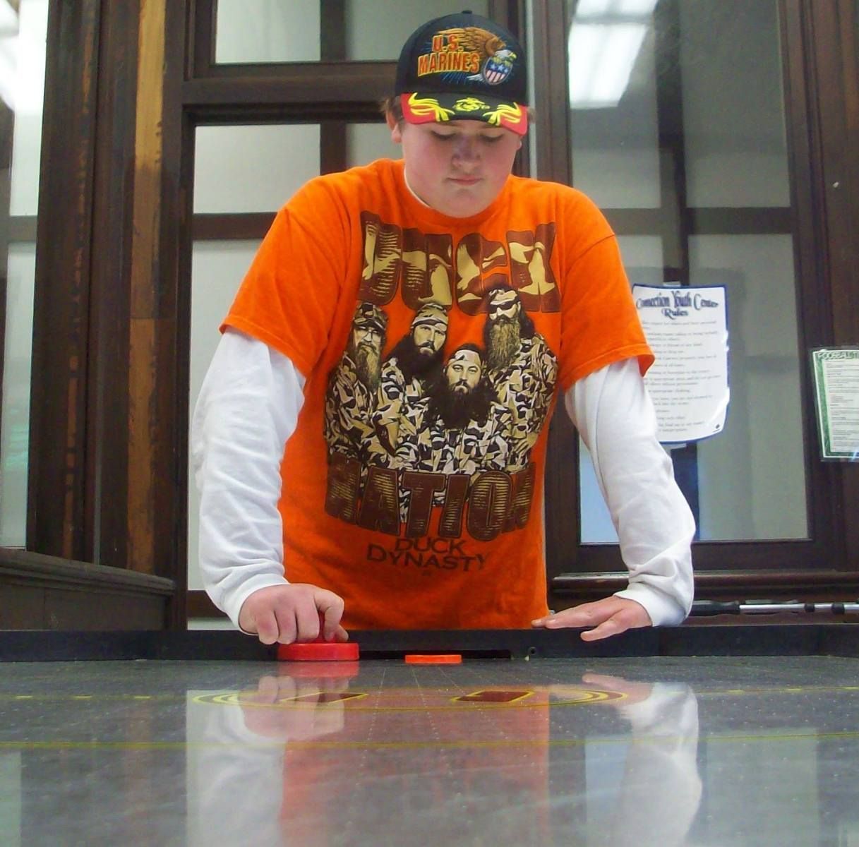 Person in orange shirt playing air hockey. They wear a hat, standing near a dark wooden structure.