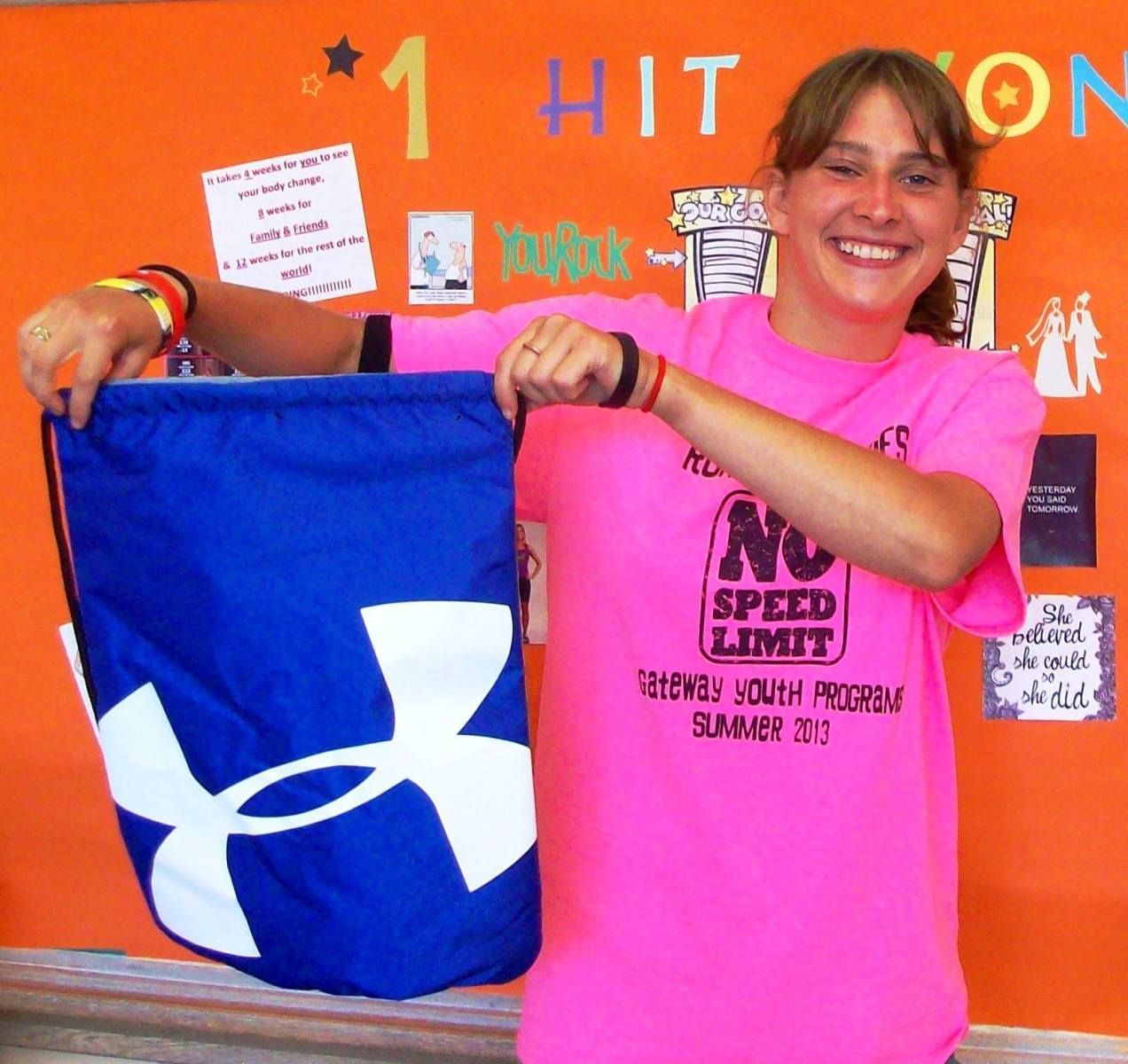 Woman in pink shirt holding a blue Under Armour drawstring bag in front of an orange bulletin board.