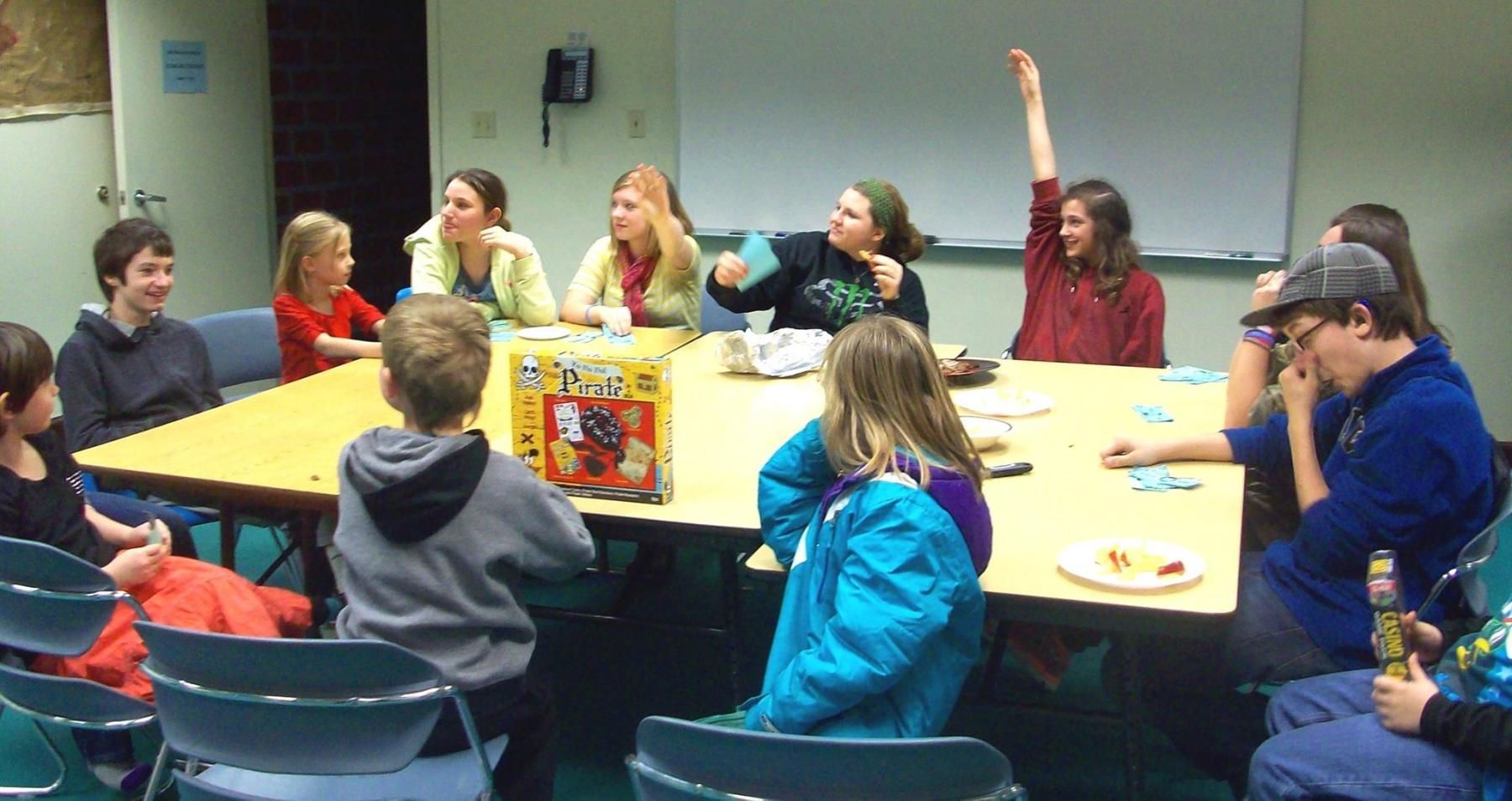 People playing a board game around a table in a room. One person has their arm raised.