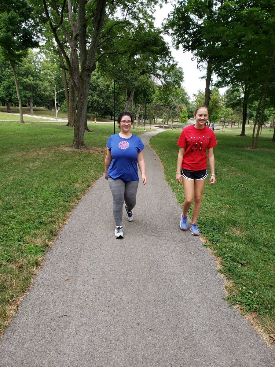 Two people walking on a paved path in a park with green grass and trees.