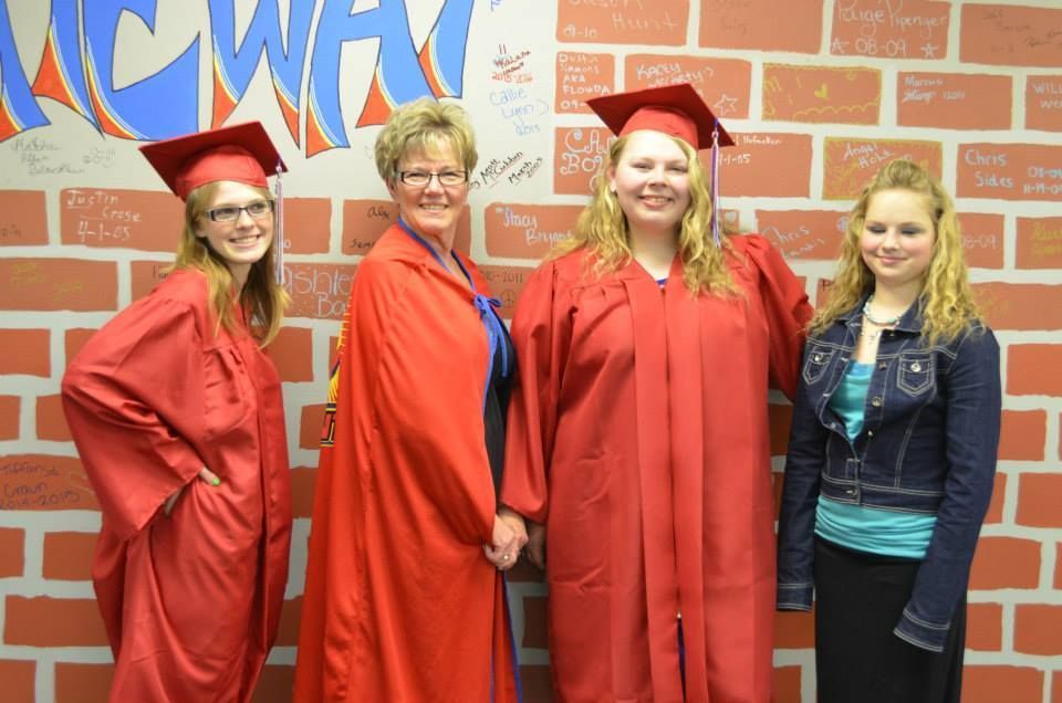 Four people stand in front of a brick wall. Three in red graduation gowns, one in a denim jacket.