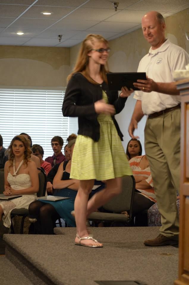 Woman in a green dress and glasses receives award from a man on stage; audience seated.