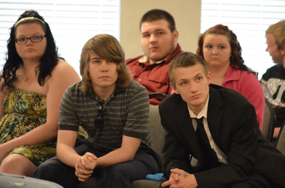 Group of young people sitting and looking forward. Some appear attentive, one wearing a suit, others in casual clothing.