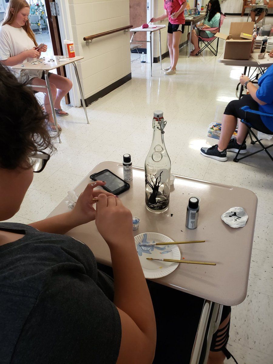 Person painting a bottle in a room with other people. Paints, brushes, and a phone are on the table.