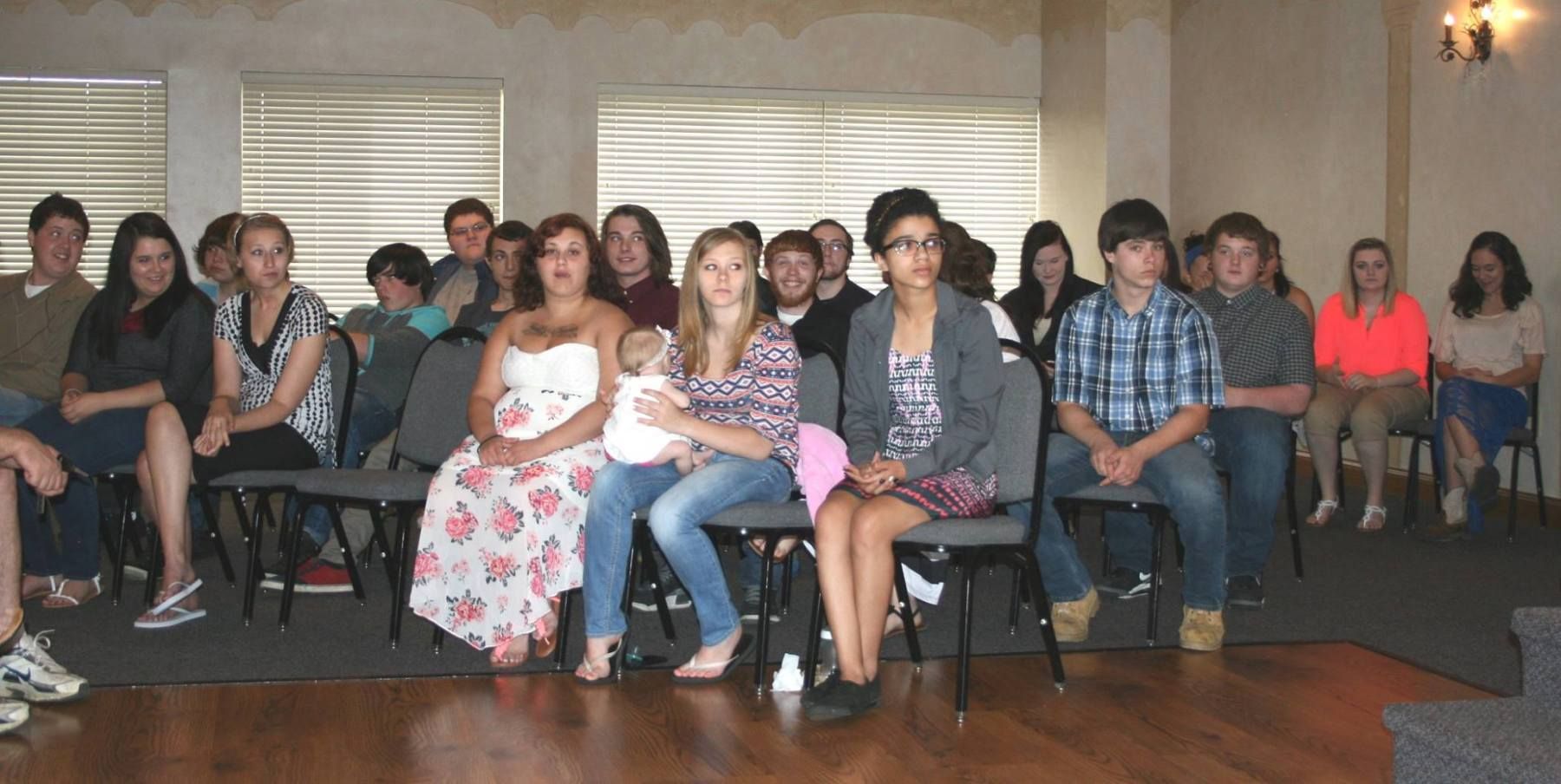 Group of people sitting in chairs in a room, some are looking at the camera.